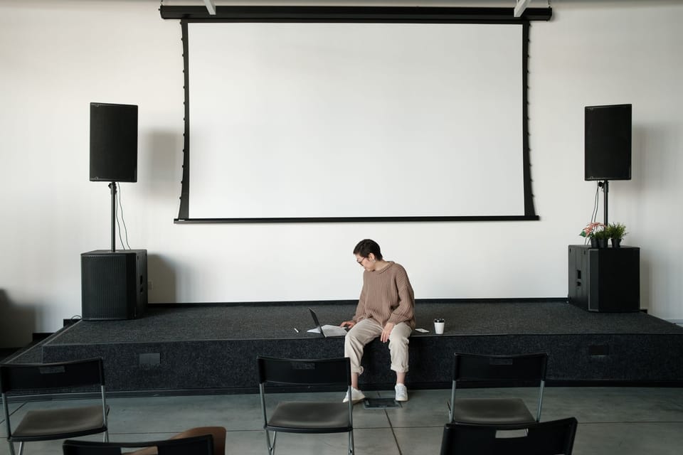 A person sitting on the edge of a stage with a laptop, under a blank projection screen between two speakers