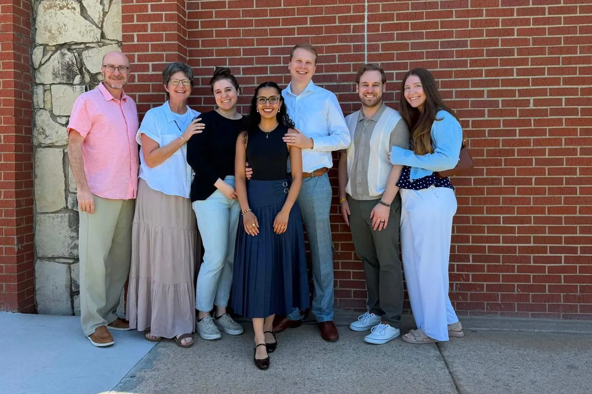 The Hamilton family, seven people standing together in front of a red brick wall on a sunny day
